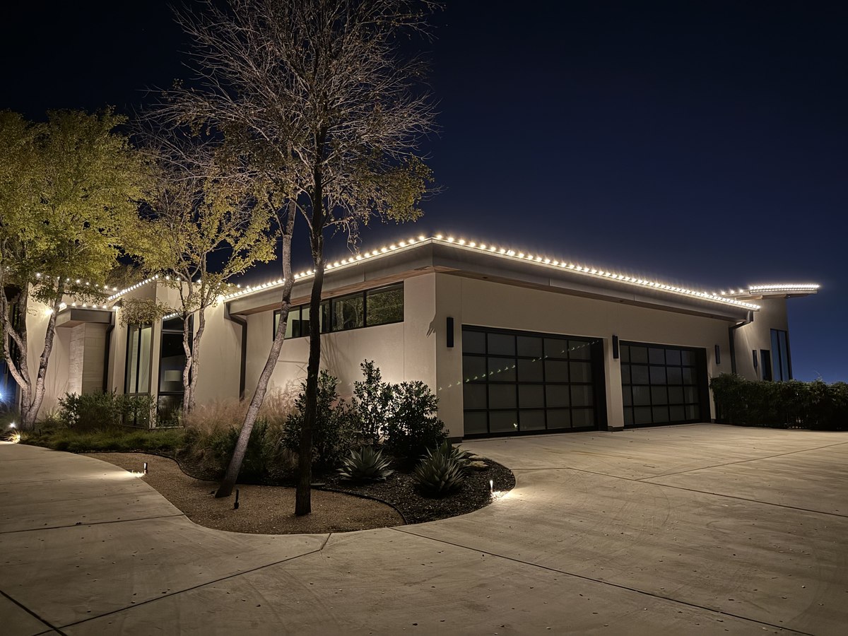 Contemporary flat-roof home with a crisp line of warm white lights along the eave and a lit tree beside the driveway