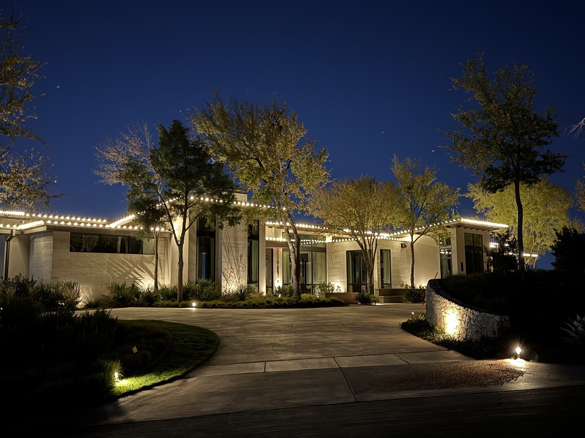 Single-story modern home lit with warm white roofline lights at blue hour, with uplit trees along the driveway
