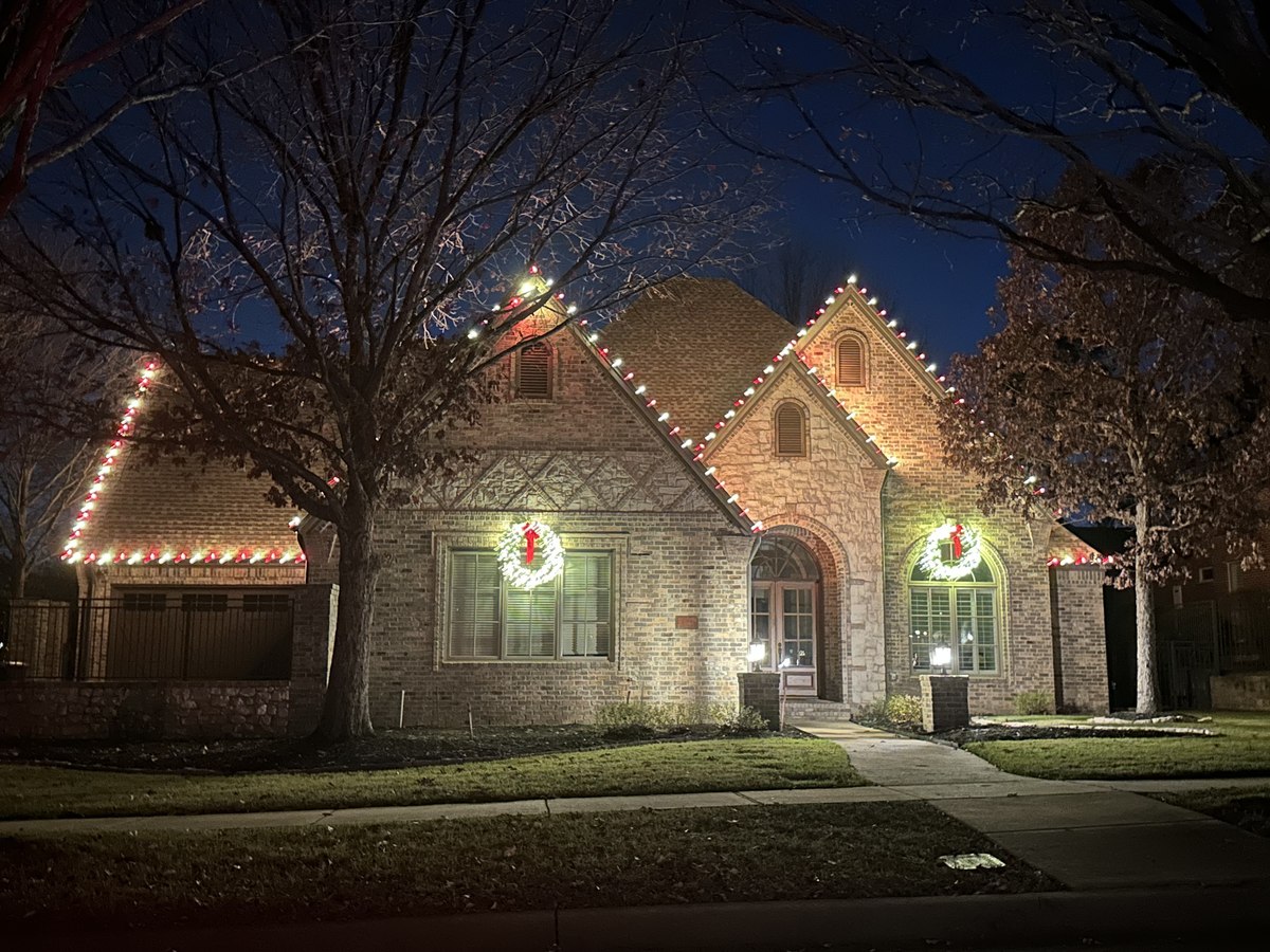 Traditional brick home with warm white roofline lights and lit wreaths on the gables, framed by bare winter trees