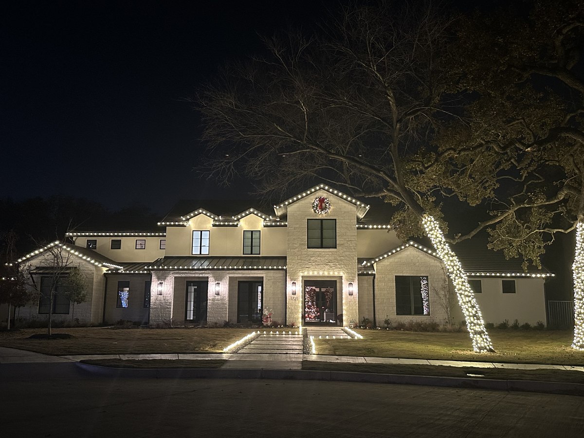 Two-story stone home with warm white roofline lighting and two tall trees wrapped in lights beside the driveway